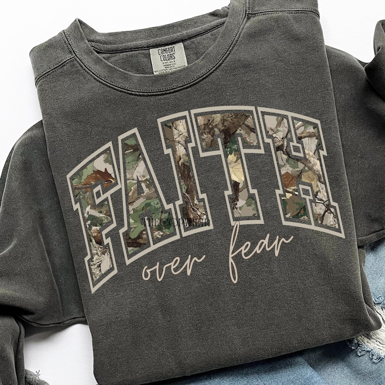 May include: Dark gray sweatshirt with the words "FAITH" in a block letter design filled with a camouflage pattern. The words "over fear" are written in a cursive font below the word "FAITH".