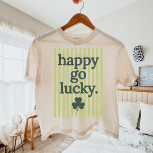 May include: Cream-colored t-shirt with the words "happy go lucky" in dark green, set against a light green striped background. A green shamrock is below the text. The shirt is hanging on a wooden hanger.