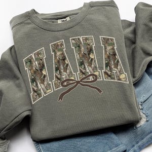 May include: An olive green sweatshirt with the word "NANA" in white outlined, camouflage-filled letters. A brown bow graphic is centered below the word. The sweatshirt is folded on top of a pair of blue denim shorts.