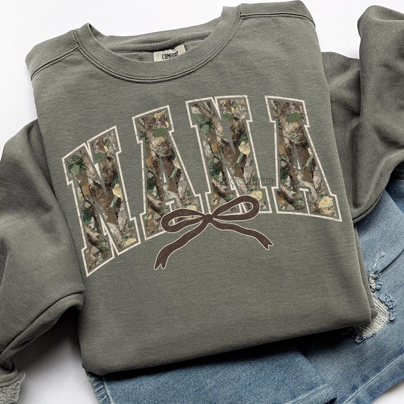 May include: An olive green sweatshirt with the word "NANA" in white outlined, camouflage-filled letters. A brown bow graphic is centered below the word. The sweatshirt is folded on top of a pair of blue denim shorts.