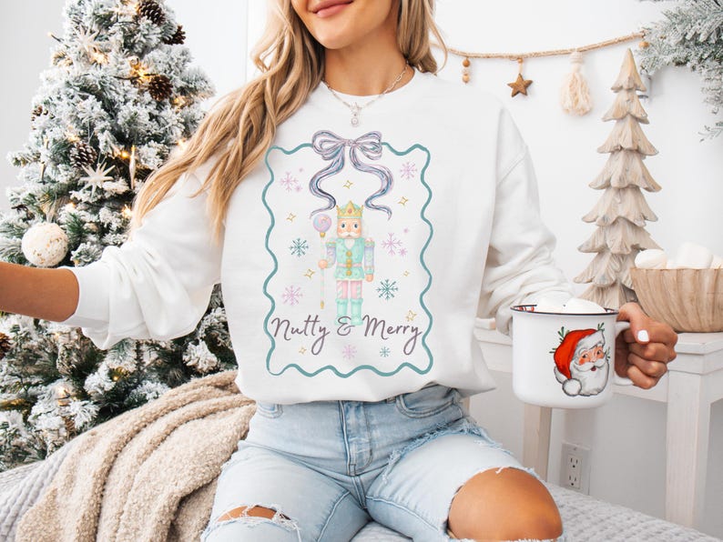 K&ouml;nnte beinhalten: Wei&szlig;es Sweatshirt mit einer Aquarell-Illustration eines Nussknackers, Schneeflocken und dem Text "Nutty & Merry". Das Design ist von einem t&uuml;rkisfarbenen Rahmen umgeben. Eine wei&szlig;e Tasse mit einer Weihnachtsmann-Grafik ist ebenfalls zu sehen.