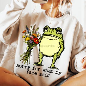 May include: Cream-colored sweatshirt featuring a cartoon frog holding a bouquet of colorful flowers. The frog is green with a yellow belly. Text on the sweatshirt reads "sorry for what my face said."
