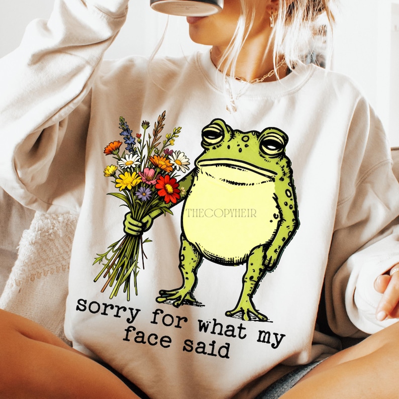 May include: Cream-colored sweatshirt featuring a cartoon frog holding a bouquet of colorful flowers. The frog is green with a yellow belly. Text on the sweatshirt reads "sorry for what my face said."