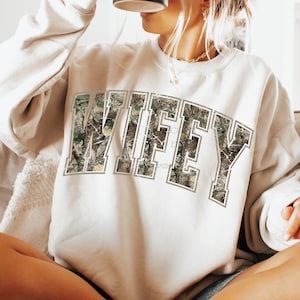 May include: Cream-colored sweatshirt with the word "WIFEY" in large, block letters across the chest. The letters are filled with a camouflage pattern and outlined in a light gray. The sweatshirt has long sleeves and a crew neck.