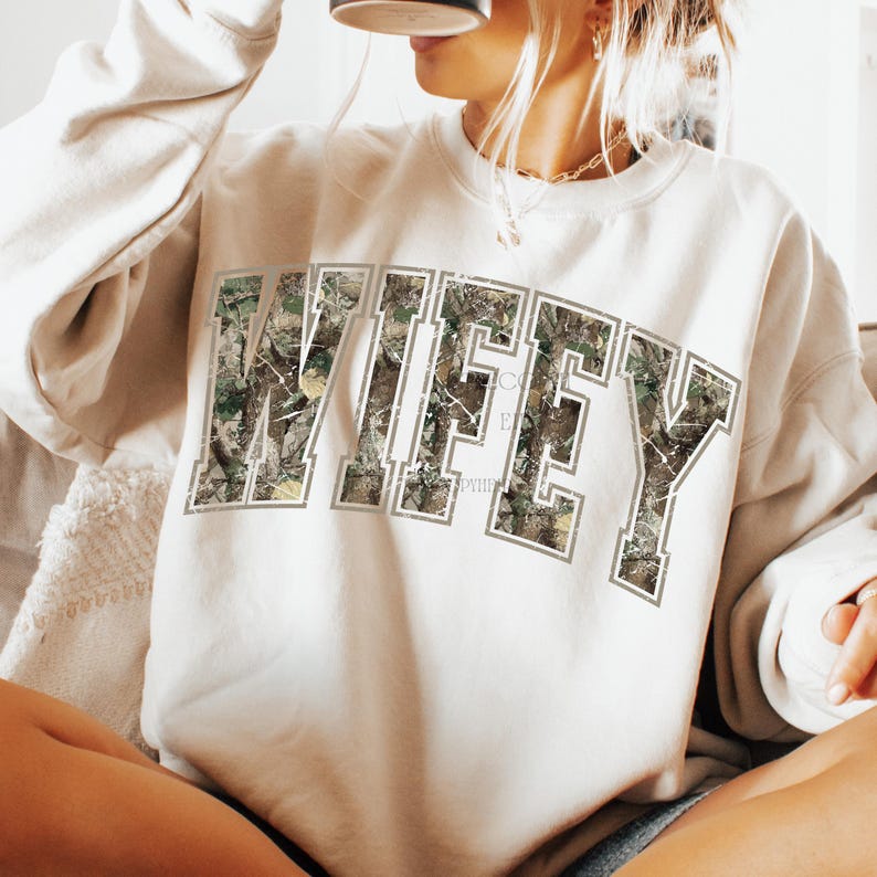 May include: Cream-colored sweatshirt with the word "WIFEY" in large, block letters across the chest. The letters are filled with a camouflage pattern and outlined in a light gray. The sweatshirt has long sleeves and a crew neck.