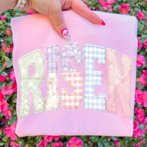 May include: A light pink sweatshirt with the word "RISEN" in patchwork letters. The letters are in various patterns, including floral, gingham, and solid colors. The sweatshirt has a crew neck and long sleeves.