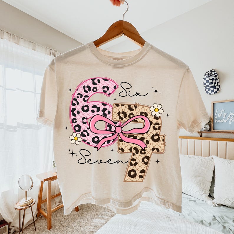 Pode incluir: Uma camiseta creme com os n&uacute;meros "6" e "7" em um design l&uacute;dico. O "6" &eacute; rosa com estampa de leopardo, e o "7" &eacute; bege com estampa de leopardo. Um la&ccedil;o rosa e margaridas acentuam os n&uacute;meros. As palavras "Six" e "Seven" est&atilde;o escritas em it&aacute;lico.
