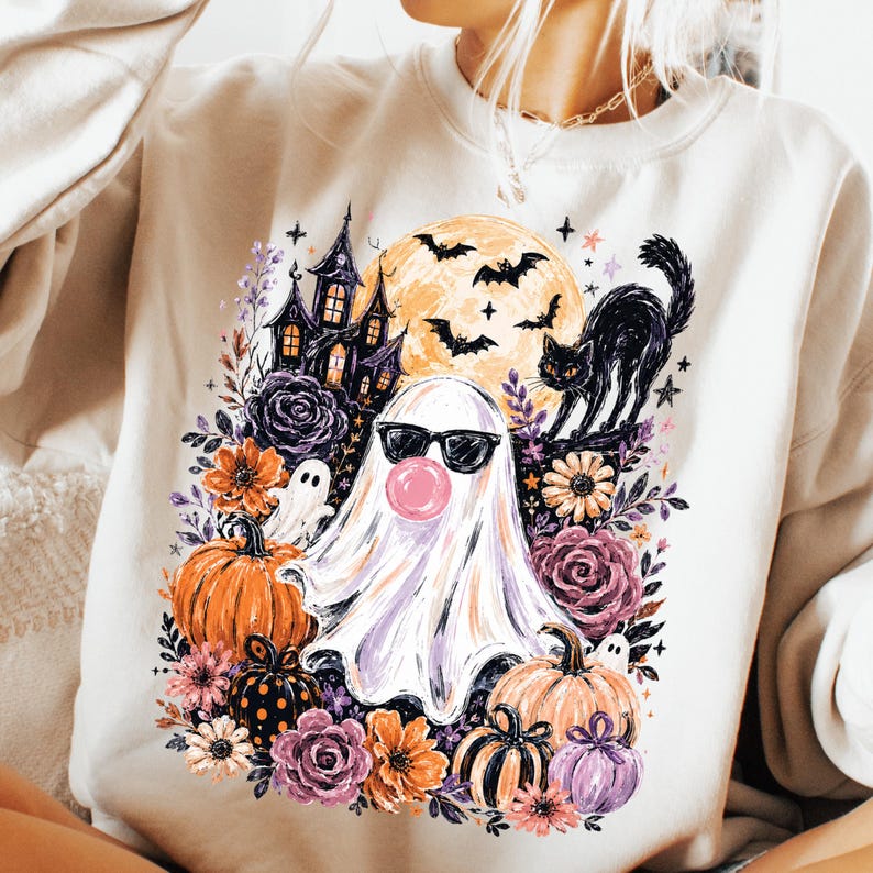 May include: A beige crewneck sweatshirt featuring a graphic print of a white ghost wearing black sunglasses and blowing a pink bubble. The design includes a haunted house, a black cat, bats, a full moon, orange pumpkins, and purple flowers.