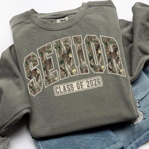 May include: A sage green sweatshirt with the word "SENIOR" in large, white, block letters filled with a camouflage pattern. Below the word "SENIOR" is the text "CLASS OF 2026" in a white rectangular box. The sweatshirt is folded on top of denim shorts.
