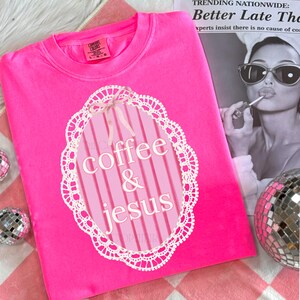 May include: A bright pink crew neck sweatshirt with the words "coffee & jesus" in a white, decorative font inside a pink and white oval design. The design is framed by a white lace-like border. The sweatshirt is on a pink and white checkered surface.