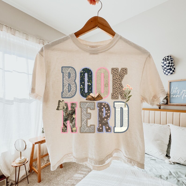 May include: Cream-colored t-shirt with the words "BOOK NERD" in a collage of patterns and textures. The letters feature floral, leopard print, and book page designs, with a butterfly and flowers. The shirt is hanging on a wooden hanger.