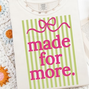 Puede incluir: Camiseta color crema con el texto "made for more" en rosa brillante. El texto est&aacute; sobre un fondo de rayas verdes verticales y un dise&ntilde;o de lazo rosa. Un delicado collar de mariposas est&aacute; drapeado cerca del cuello.