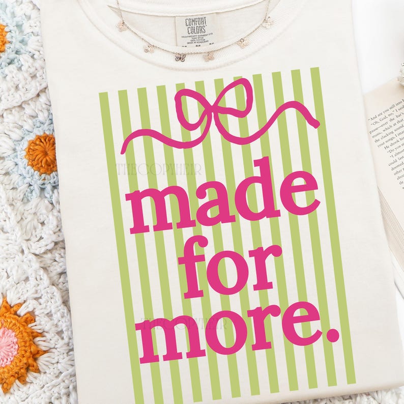 Puede incluir: Camiseta color crema con el texto "made for more" en rosa brillante. El texto est&aacute; sobre un fondo de rayas verdes verticales y un dise&ntilde;o de lazo rosa. Un delicado collar de mariposas est&aacute; drapeado cerca del cuello.