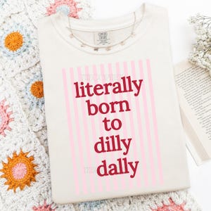 Puede incluir: Camiseta color crema con la frase "literally born to dilly dally" en rojo y rayas rosas. Un delicado collar plateado con peque&ntilde;os dijes descansa sobre la camiseta. La camiseta est&aacute; sobre una superficie blanca, con una manta de ganchillo.