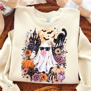 May include: A cream-coloured crewneck sweatshirt featuring a graphic print of a ghost wearing black sunglasses and blowing a pink bubble. The design includes a haunted house, a black cat, bats, a full moon, pumpkins, and various colourful flowers.