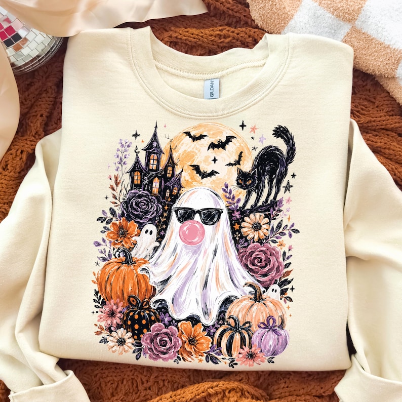 May include: A cream-coloured crewneck sweatshirt featuring a graphic print of a ghost wearing black sunglasses and blowing a pink bubble. The design includes a haunted house, a black cat, bats, a full moon, pumpkins, and various colourful flowers.