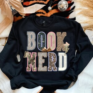 May include: Black sweatshirt with the words "BOOK NERD" in a colorful, layered design. The letters incorporate floral, leopard print, and book page patterns. A small open book graphic is between the "O" and "K".