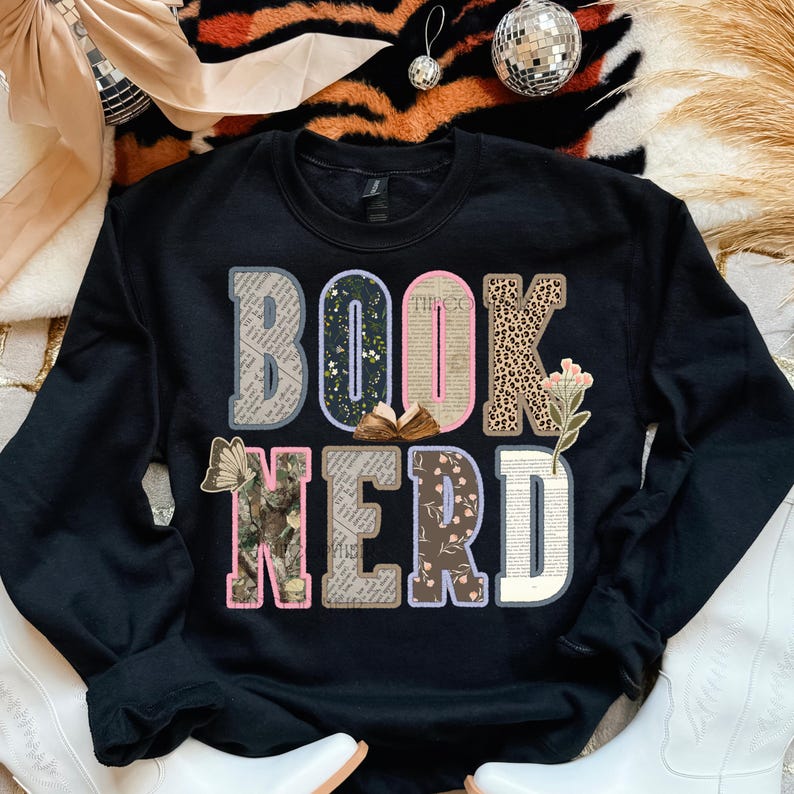 May include: Black sweatshirt with the words "BOOK NERD" in a colorful, layered design. The letters incorporate floral, leopard print, and book page patterns. A small open book graphic is between the "O" and "K".