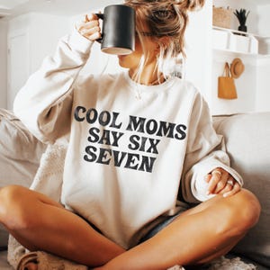 May include: A cream-colored sweatshirt with the text "COOL MOMS SAY SIX SEVEN" in black. The sweatshirt is being held by a person with a black mug. The person is sitting on a couch.
