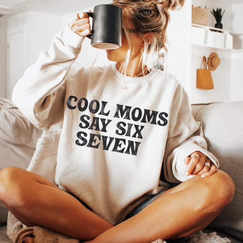 May include: A cream-colored sweatshirt with the text "COOL MOMS SAY SIX SEVEN" in black. The sweatshirt is being held by a person with a black mug. The person is sitting on a couch.