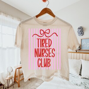 May include: A beige t-shirt with the words "TIRED NURSES CLUB" in red, set against a pink striped background. A red bow design is above the text. The shirt is hanging on a wooden hanger.