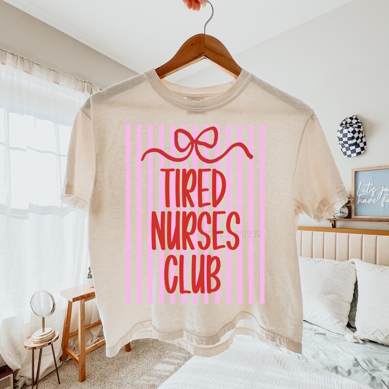 May include: A beige t-shirt with the words "TIRED NURSES CLUB" in red, set against a pink striped background. A red bow design is above the text. The shirt is hanging on a wooden hanger.