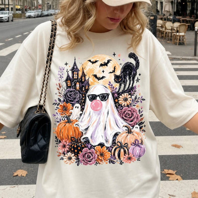 May include: A cream-coloured t-shirt featuring a graphic print of a ghost wearing sunglasses and blowing a pink bubble, surrounded by a haunted house, a black cat, bats, a full moon, pumpkins, and various colourful flowers in shades of orange and purple.