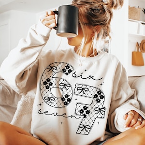 May include: A cream-colored sweatshirt with the numbers "67" in a black lace design, adorned with bows and floral accents. The word "six" and "seven" are written in cursive above and below the numbers. A black mug is held up to the face.