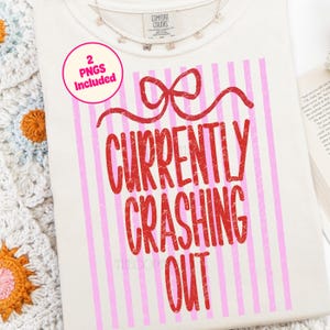 以下が含まれることがあります： 赤いリボンと「CURRENTLY CRASHING OUT」の文字が赤色でプリントされたクリーム色のTシャツ。デザインにはピンクの縦縞が含まれています。蝶のチャームが付いた小さなネックレスが見えます。