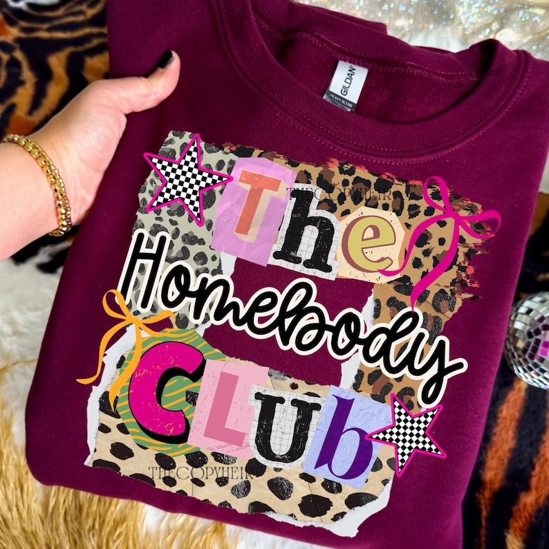 May include: Burgundy sweatshirt with the text "The Homebody Club" in a collage style. The design includes leopard print, stars, and bows. The sweatshirt is a Gildan brand.