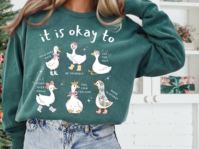 Pode incluir: Camisola verde-azulado com a frase "it is okay to" acima de gansos de desenho animado com mensagens encorajadoras como "Have hard days" e "Make mistakes". Os gansos usam acess&oacute;rios como botas e la&ccedil;os.