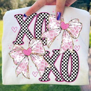 May include: White t-shirt featuring the letters "XO" in a black and white checkered pattern, with pink outlines. Two large bows with pink floral patterns and glittery pink hearts are also featured. Small pink hearts and stars complete the design.