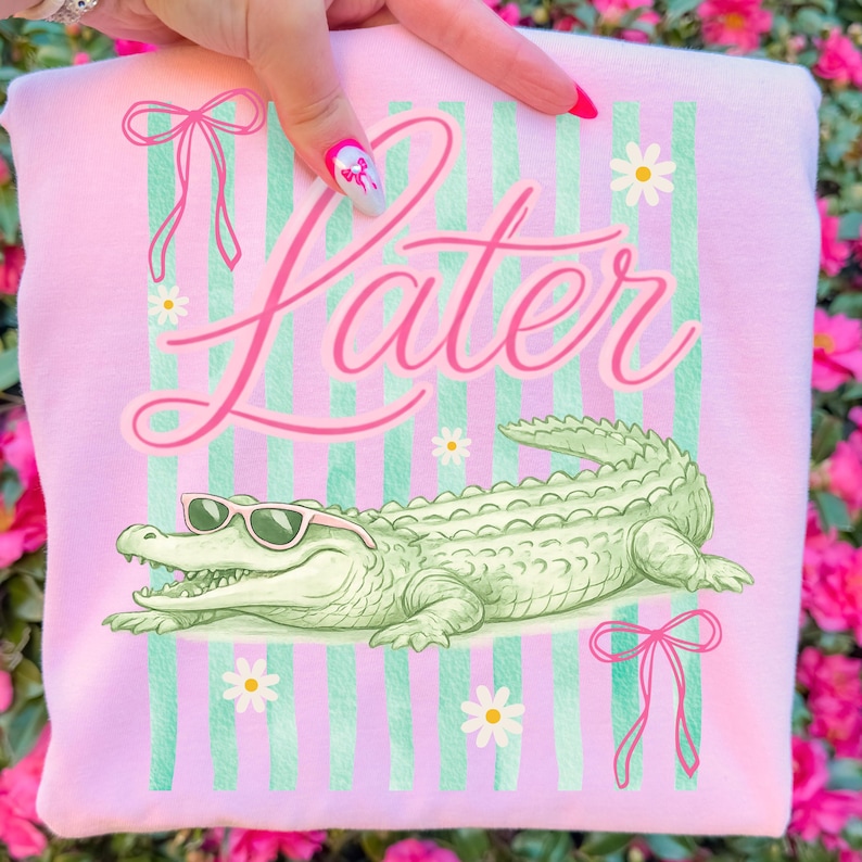 K&ouml;nnte beinhalten: Rosa Sweatshirt mit dem Wort "Later" in Schreibschrift. Das Design umfasst ein gr&uuml;nes Alligator mit Sonnenbrille, gr&uuml;n-wei&szlig;e Streifen, G&auml;nsebl&uuml;mchen und rosa Schleifen. Ein lustiges, l&auml;ssiges Design.