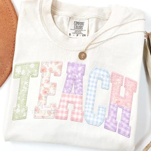 Puede incluir: Camiseta color crema con la palabra "TEACH" en letras de patchwork pastel. Las letras presentan patrones florales, de cuadros y lisos. Un collar de oro con un colgante descansa sobre la camiseta. La camiseta tiene una etiqueta Comfort Colors.