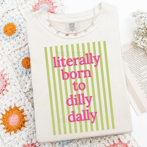Puede incluir: Camiseta color crema con la frase "literally born to dilly dally" en letras rosas, sobre un fondo de rayas verticales verdes. Un delicado collar de mariposas descansa sobre la camiseta. La camiseta es de Comfort Colors.