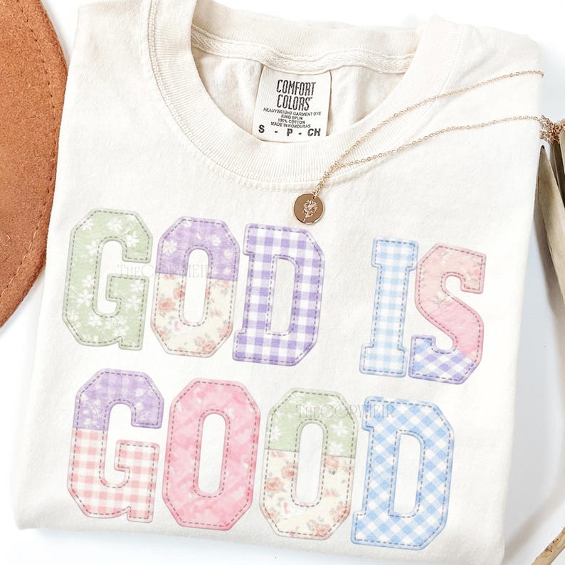 May include: Cream-colored t-shirt with the phrase "GOD IS GOOD" in patchwork letters. The letters feature floral and gingham patterns in pastel colors. A gold necklace with a pendant rests on the shirt.