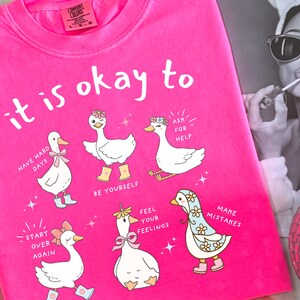 Pode incluir: Camiseta rosa choque com a frase "it is okay to" e ilustra&ccedil;&otilde;es de gansos de desenho animado com mensagens inspiradoras. Os gansos usam acess&oacute;rios como la&ccedil;os e botas. Uma foto em preto e branco est&aacute; no fundo.