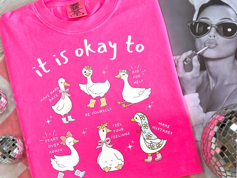 Pode incluir: Camiseta rosa choque com a frase "it is okay to" e ilustra&ccedil;&otilde;es de gansos de desenho animado com mensagens inspiradoras. Os gansos usam acess&oacute;rios como la&ccedil;os e botas. Uma foto em preto e branco est&aacute; no fundo.