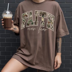 May include: Brown t-shirt with the words "FAITH over fear" in a white, arched font. The letters of "FAITH" are filled with a camouflage pattern. The shirt is a loose fit, with short sleeves.