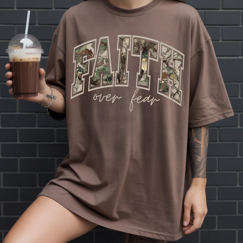 May include: Brown t-shirt with the words "FAITH over fear" in a white, arched font. The letters of "FAITH" are filled with a camouflage pattern. The shirt is a loose fit, with short sleeves.