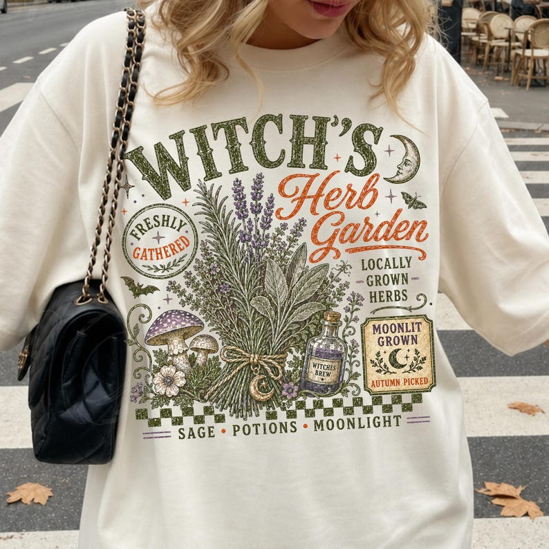 Peut inclure: Un sweat-shirt blanc cass&eacute; avec une impression graphique portant le texte "WITCH'S Herb Garden" ainsi que "FRESHLY GATHERED", "LOCALLY GROWN HERBS", "MOONLIT GROWN", "AUTUMN PICKED" et "SAGE POTIONS MOONLIGHT". Le motif inclut des herbes, des champignons et une bouteille.