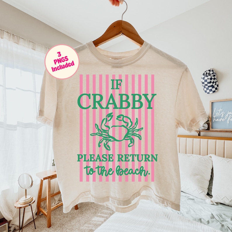 Puede incluir: Camiseta color crema con la frase "IF CRABBY PLEASE RETURN to the beach." El dise&ntilde;o incluye un cangrejo verde sobre rayas rosas y blancas. La camiseta est&aacute; colgada en una percha de madera.