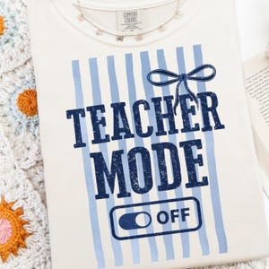 May include: Cream-colored t-shirt with blue vertical stripes and the words "TEACHER MODE OFF" in navy blue. A navy blue bow is above the words. A delicate butterfly necklace rests on the shirt.