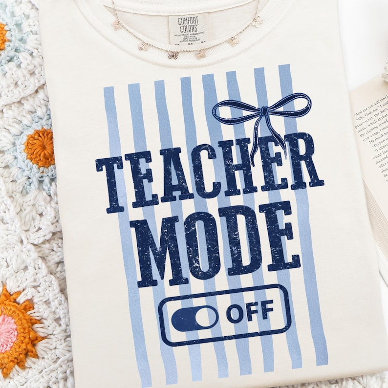 May include: Cream-colored t-shirt with blue vertical stripes and the words "TEACHER MODE OFF" in navy blue. A navy blue bow is above the words. A delicate butterfly necklace rests on the shirt.