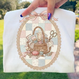 May include: White sweatshirt featuring a detailed illustration of a sewing basket. The basket is filled with spools of thread, fabric scraps, and scissors, set against a pastel patchwork background. A pink bow and oval frame complete the design.