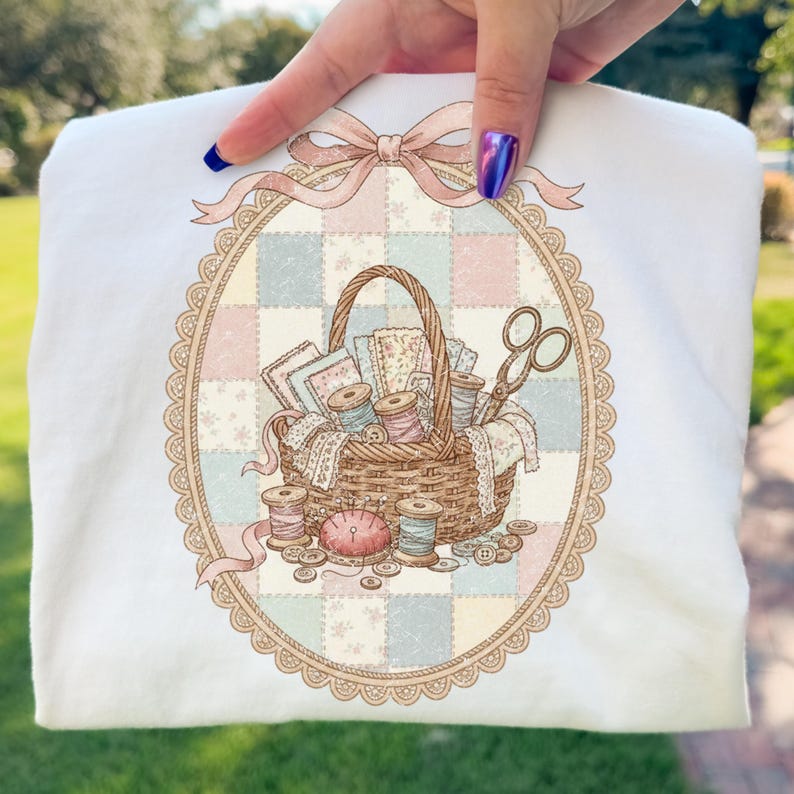 May include: White sweatshirt featuring a detailed illustration of a sewing basket. The basket is filled with spools of thread, fabric scraps, and scissors, set against a pastel patchwork background. A pink bow and oval frame complete the design.