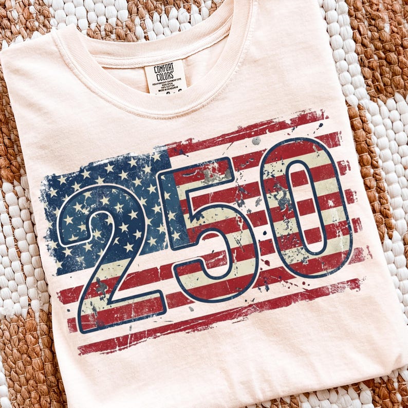 Puede incluir: Camiseta de color melocot&oacute;n claro con un dise&ntilde;o desgastado de la bandera estadounidense y el n&uacute;mero "250". El dise&ntilde;o de la bandera incorpora rayas rojas y blancas con estrellas azules. La camiseta tiene cuello redondo y mangas cortas.
