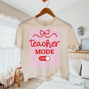 May include: Cream-colored t-shirt with a graphic that reads "teacher MODE OFF" in red, with a bow and pink vertical stripes. The shirt is hanging on a wooden hanger.