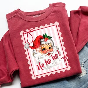May include: A maroon sweatshirt featuring a vintage Santa Claus graphic within a pink postage stamp design. The Santa illustration includes a red hat, white beard, and the words "Ho ho ho!" in a festive font. The sweatshirt is folded.