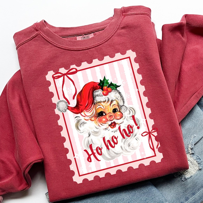 May include: A maroon sweatshirt featuring a vintage Santa Claus graphic within a pink postage stamp design. The Santa illustration includes a red hat, white beard, and the words "Ho ho ho!" in a festive font. The sweatshirt is folded.